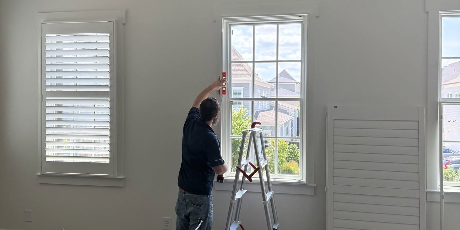 A Brentwood Blind Company team member measuring a window with a level and ladder